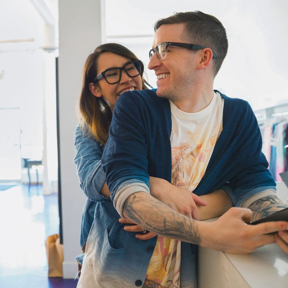 Zwei junge Leute stehen im Geschäft am Tresen, die Frau umarmt den Mann.