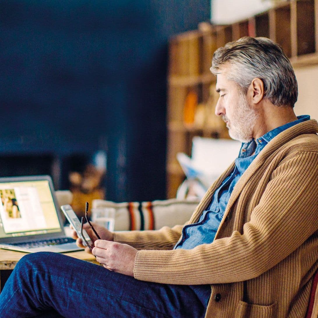 Mann sitzt vorm Laptop und schaut auf sein Handy.