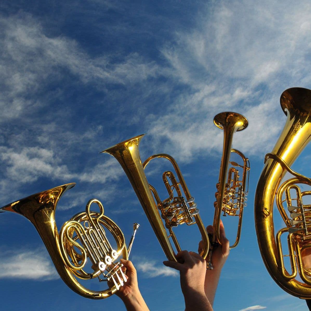 Vier Blechblasinstrumente, die vor einem blauen Himmel mit Wolken in die Höhe gehalten werden.