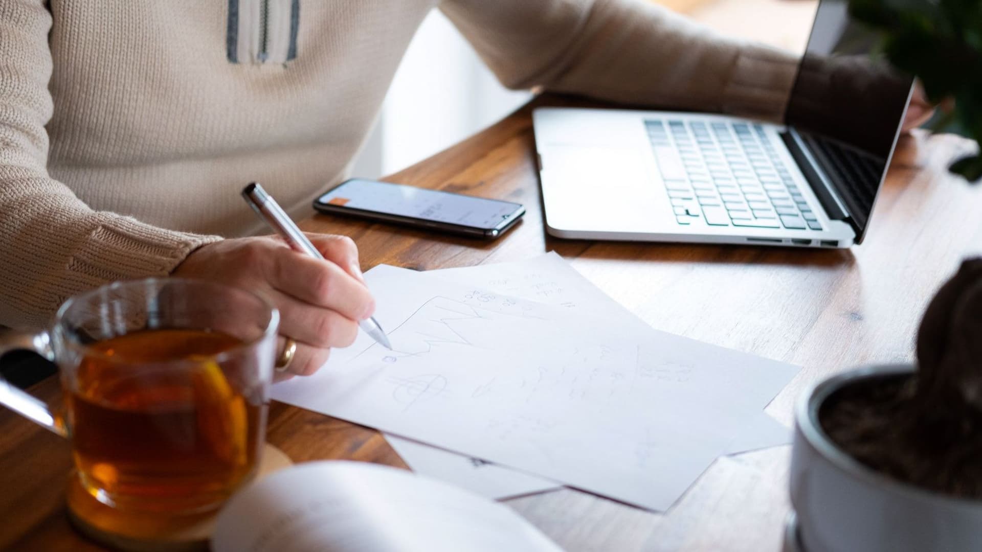 Jemand sitzt am Tisch mit Laptop, Handy und Notizen.