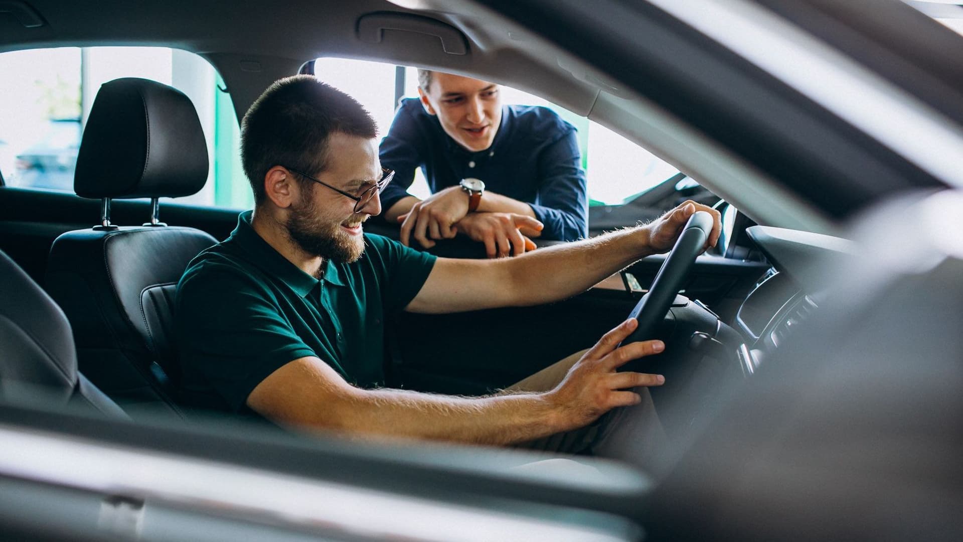 Mann sitzt im Autohaus im Auto, ein anderer steht ans Fenster gelehnt daneben.