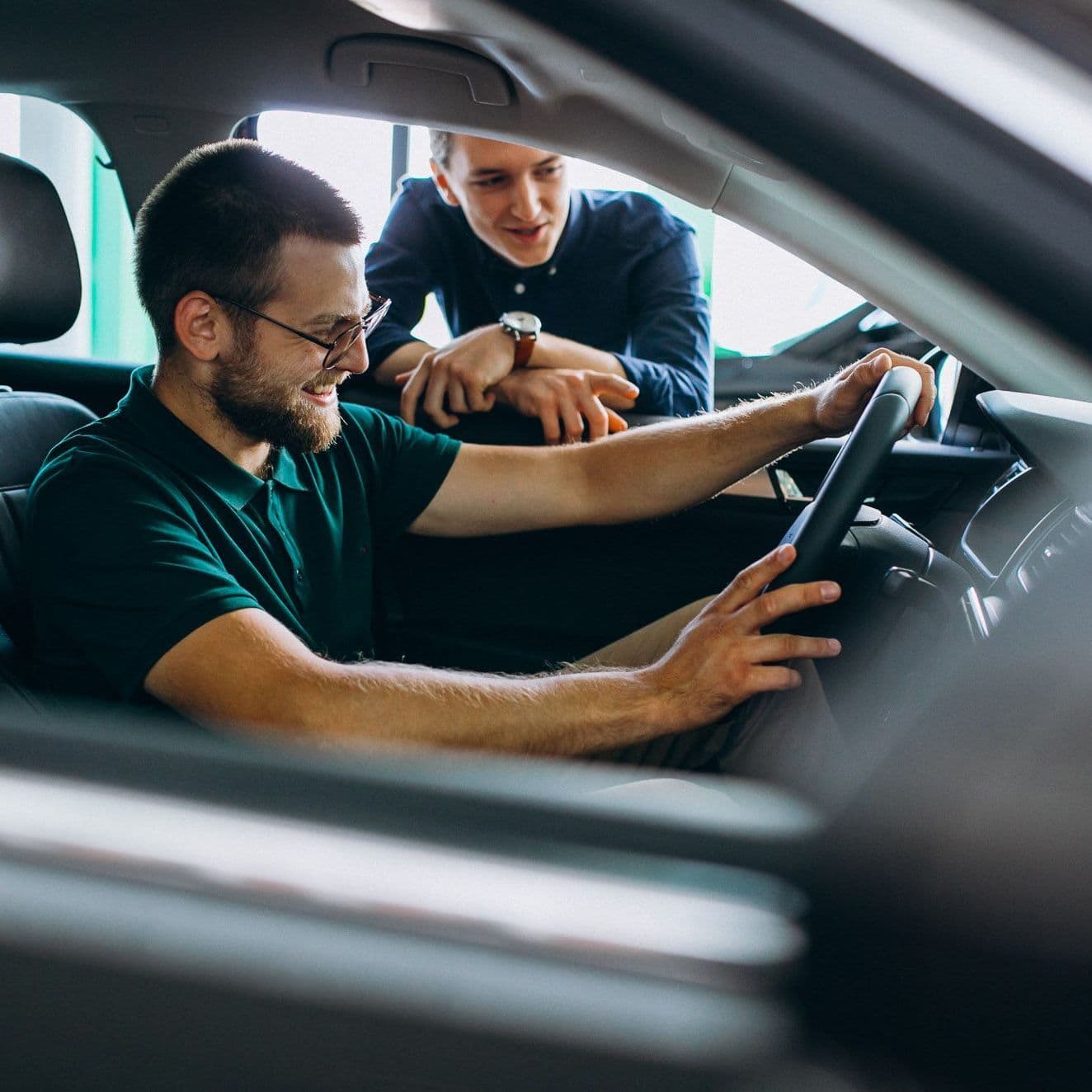 Mann sitzt im Autohaus im Auto, ein anderer steht ans Fenster gelehnt daneben.