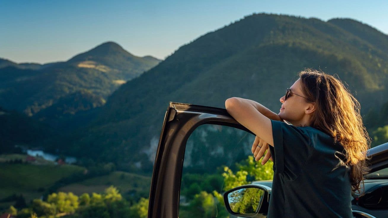 Frau lehnt sich an die Autotür und blickt in die Ferne, im Hintergrund sind Berge zu sehen.