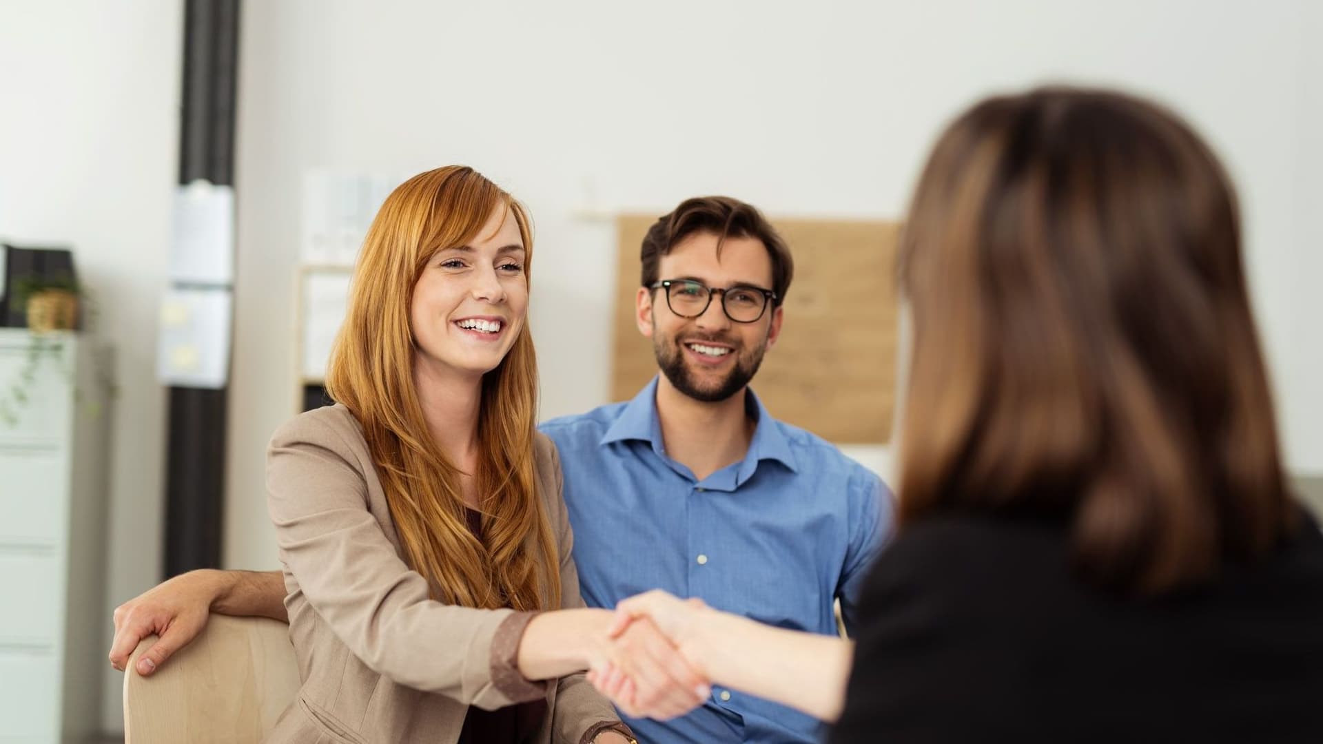 Zwei Frauen schütteln Hände, ein Mann sitzt daneben.