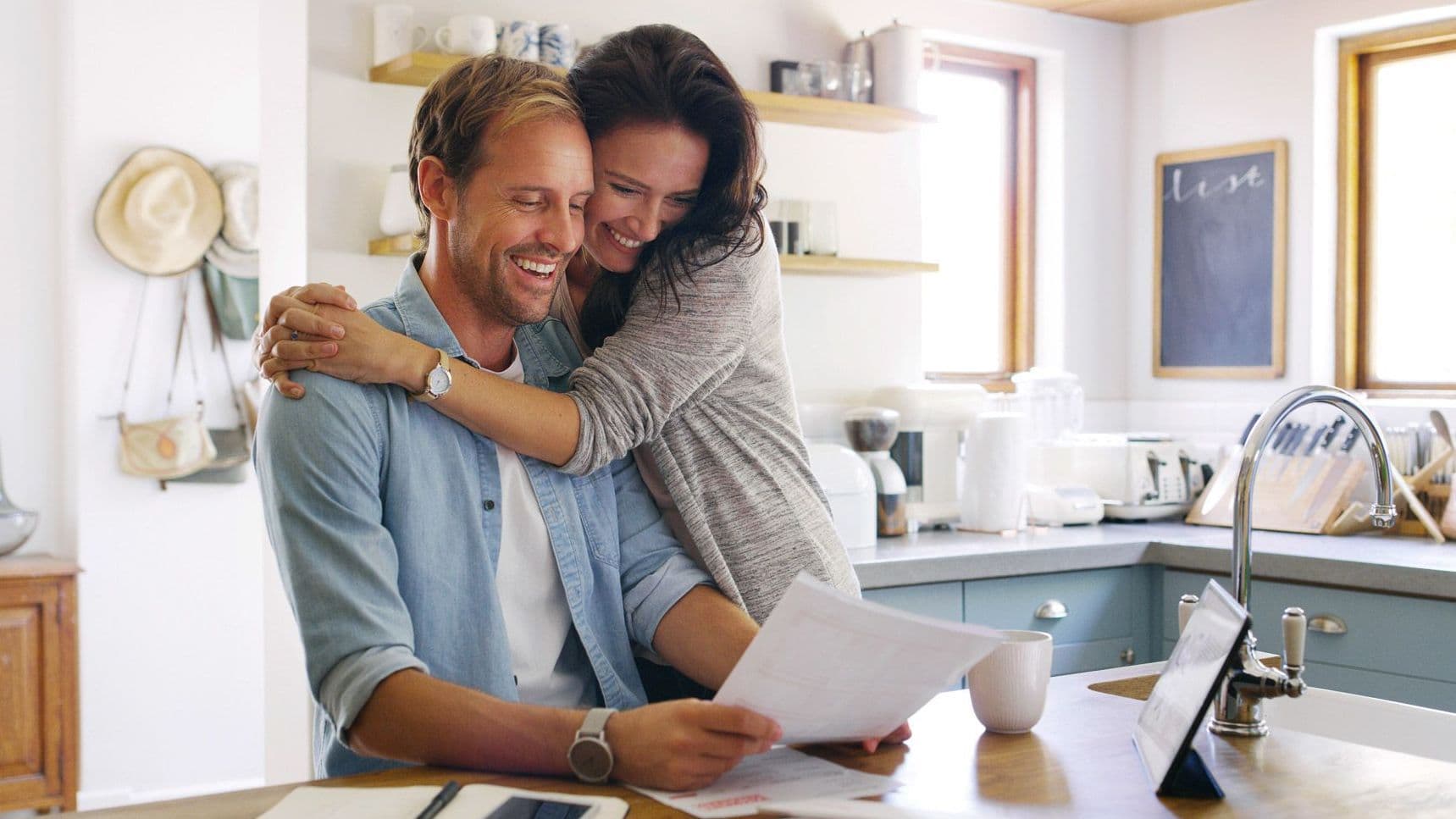Zwei Personen in der Küche, die Frau umarmt den Mann, der Mann hält Papiere in der Hand.