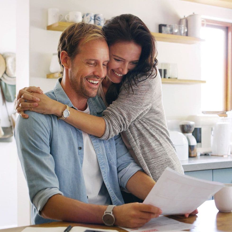 Zwei Personen in der Küche, die Frau umarmt den Mann, der Mann hält Papiere in der Hand.