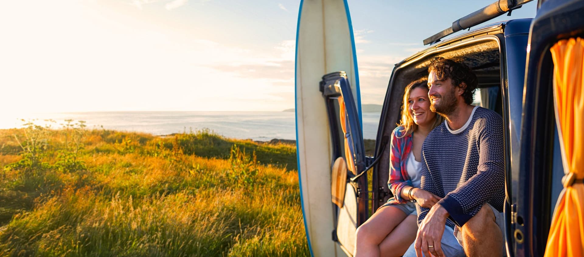 Zwei Personen sitzen im Camper nahe dem Meer, ein Surfbrett steht daneben.