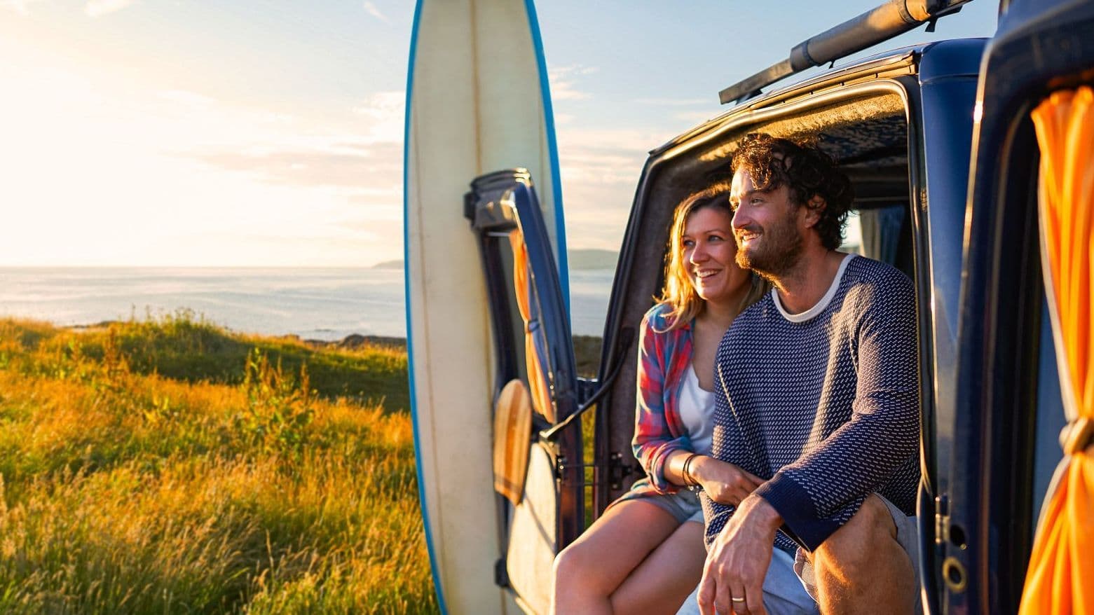 Zwei Personen sitzen im Camper nahe dem Meer, ein Surfbrett steht daneben.
