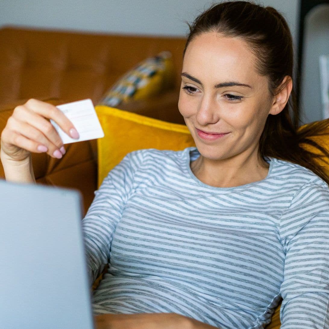 Junge Frau liegt auf dem Sofa, Laptop auf dem Schoß und Bezahlkarte in der Hand.