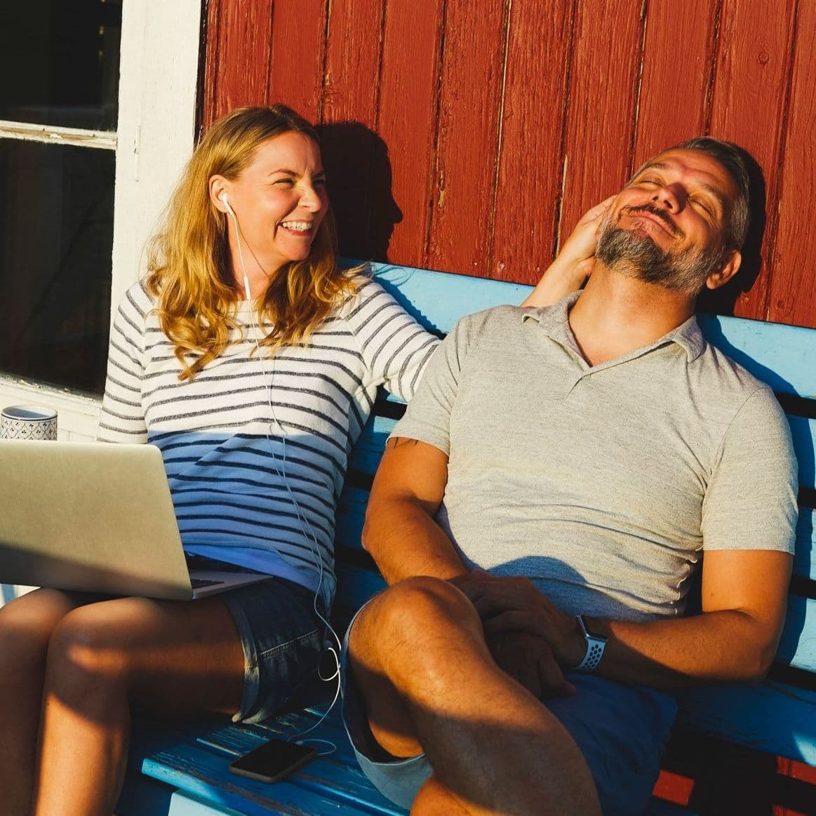 Frau und Mann sitzen vor einem Haus in der Sonne, die Frau hat einen Laptop auf dem Schoß.