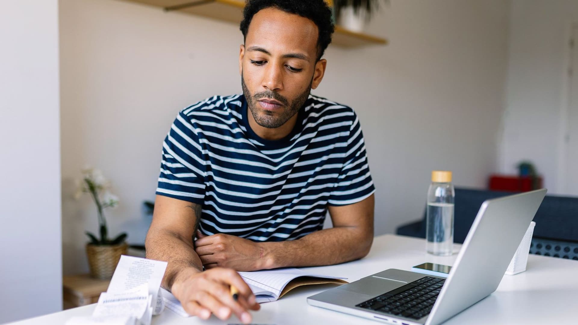 Mann sitzt am Tisch, darauf steht ein Laptop und liegen Rechnungen. Er tippt auf einem Taschenrechner.