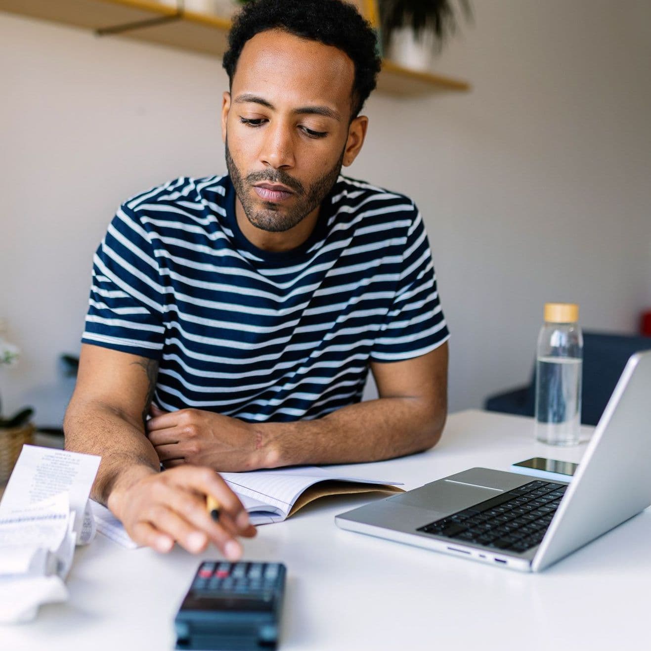 Mann sitzt am Tisch, darauf steht ein Laptop und liegen Rechnungen. Er tippt auf einem Taschenrechner.