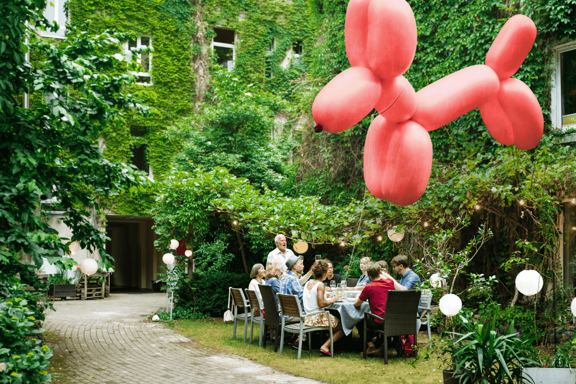 Ein pinkfarbener Ballonhund fliegt durch einen Hof, im Hintergrund sitzen Menschen beieinander.