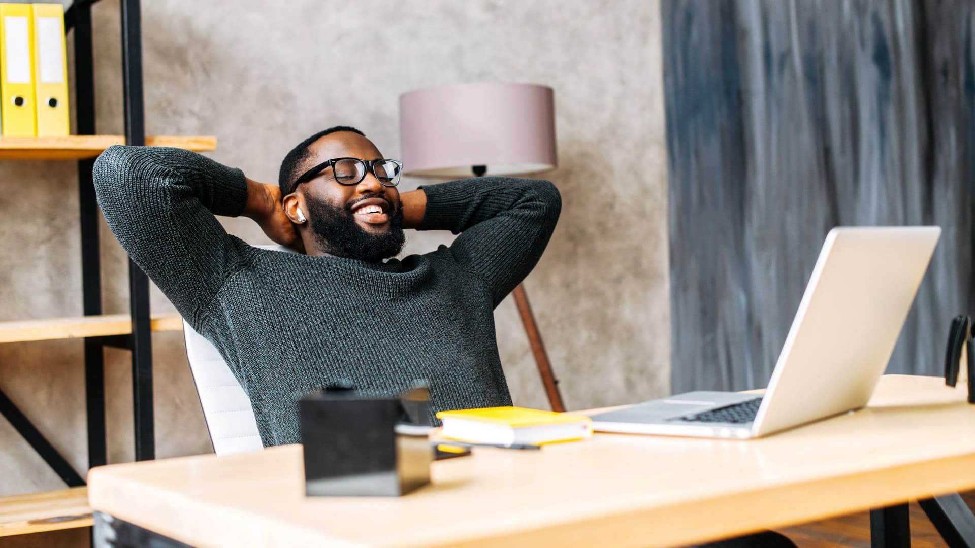 Junger Mann sitzt am Laptop, die Arme hinterm Kopf verschränkt, lächelnd.